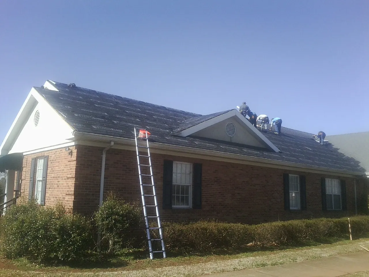Crew tearing shingles off a brick ranch roof with a ladder set — Metal Roof Repair work by our crew in Mineola